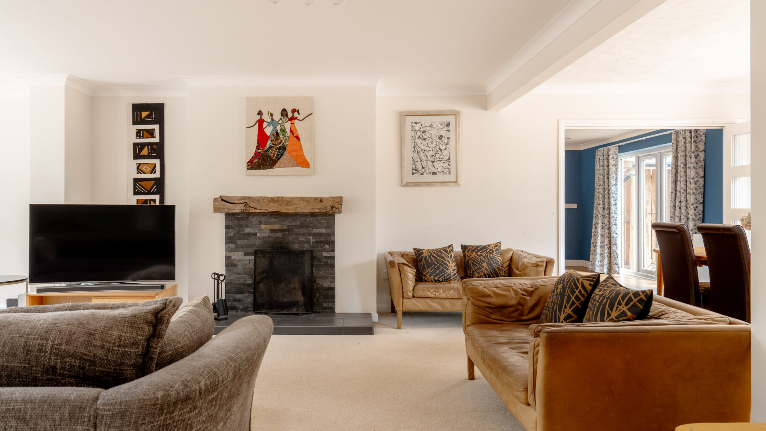 A modern living room with a fireplace, TV, and two sofas. Colorful artwork hangs above the fireplace. A dining area is visible in the background with a large window bringing in natural light.