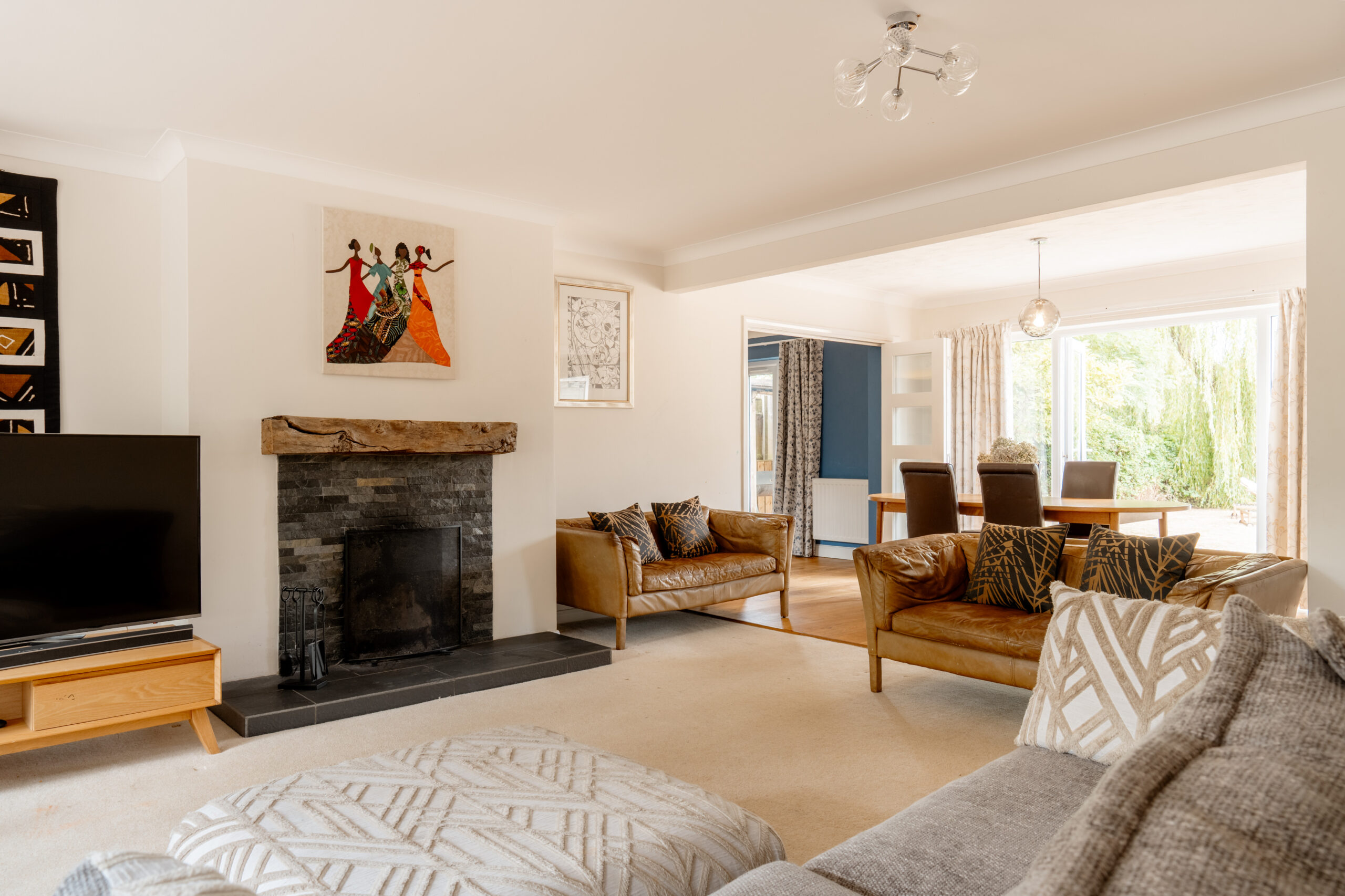 Bright living room with a fireplace, modern art above the mantle, a TV, two sofas with patterned pillows, and a dining area with a wooden table and chairs seen through a wide opening. Large windows bring in natural light.