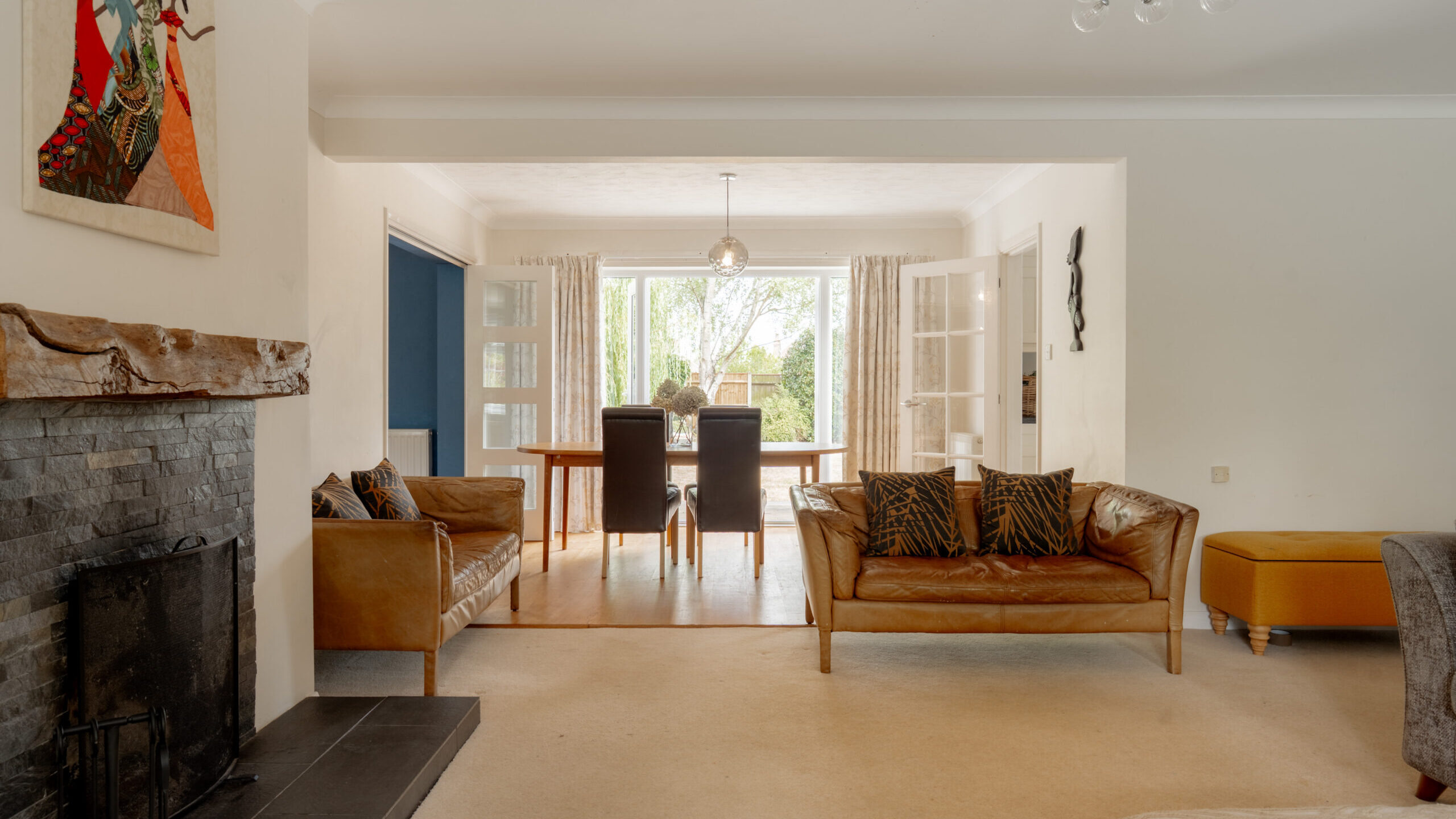 A bright living room with tan sofas, a stone fireplace, animal print cushions, art on the wall, and a view into a dining area with a table and four chairs by glass doors leading to a garden.