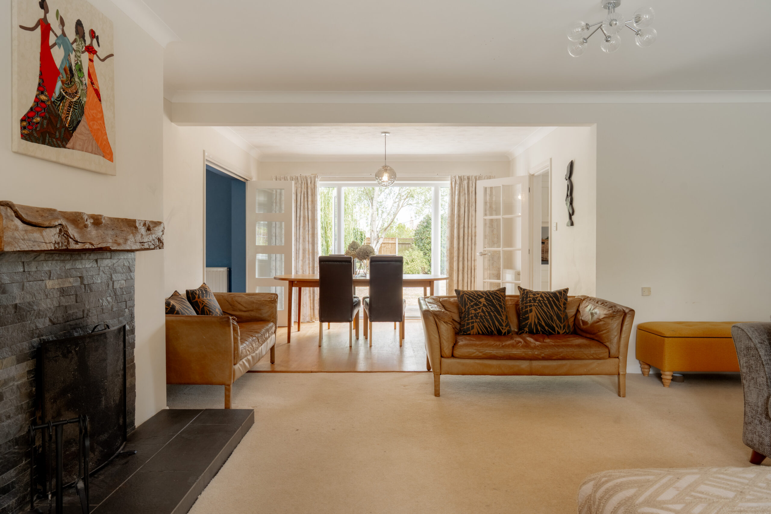 A bright living room with tan sofas, a stone fireplace, animal print cushions, art on the wall, and a view into a dining area with a table and four chairs by glass doors leading to a garden.