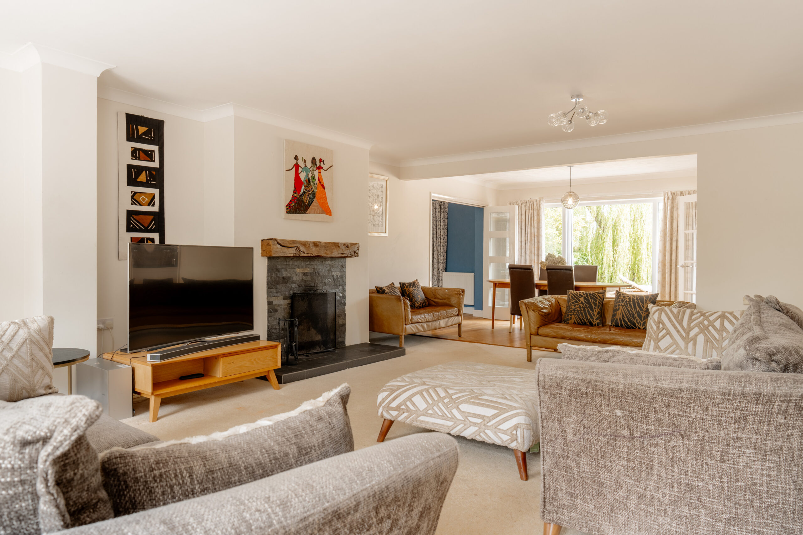 A spacious living room with beige walls, gray sofas, a modern fireplace, TV on a wooden stand, artwork above the mantel, and a view into a dining area with large windows letting in natural light.