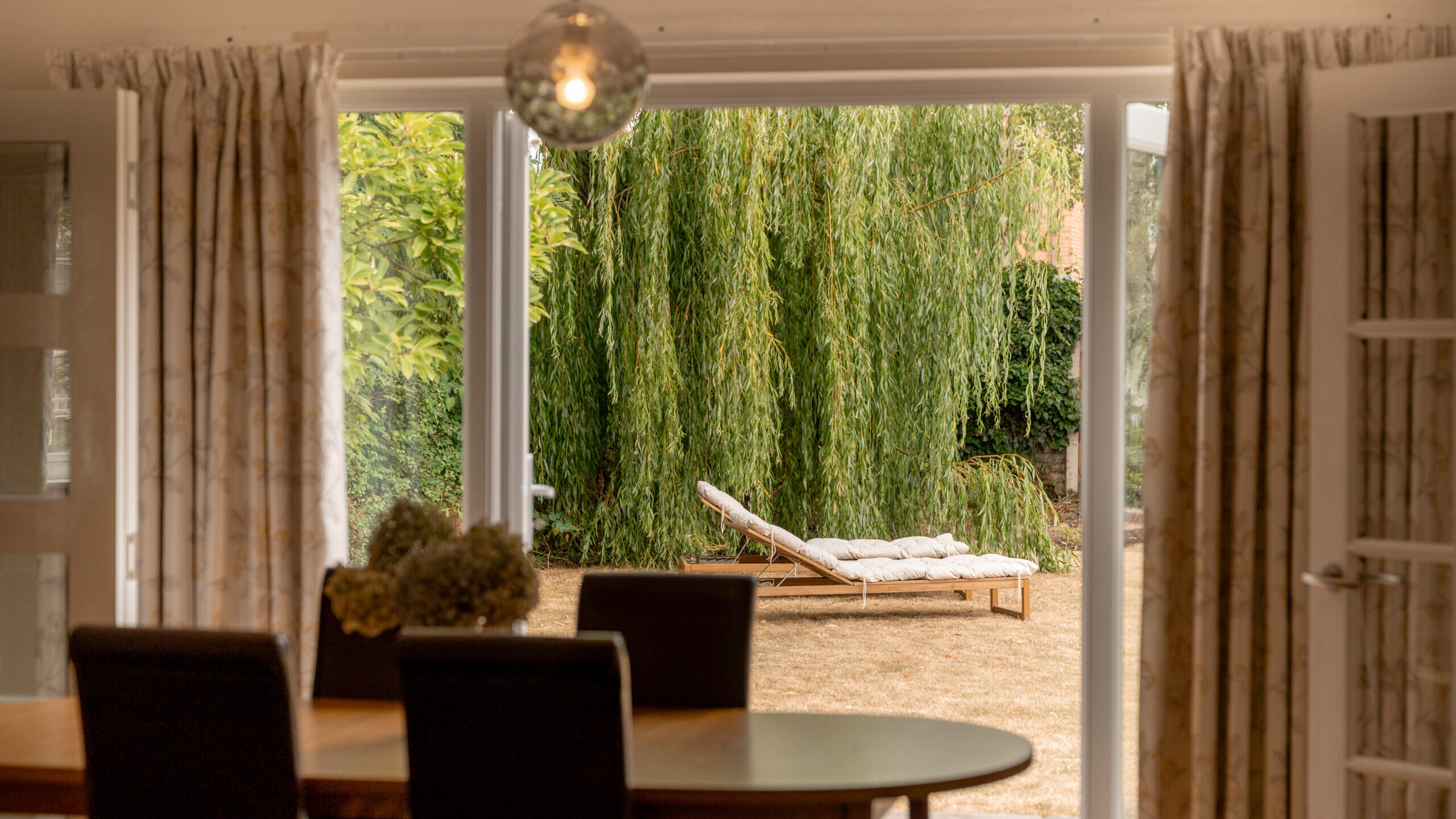Dining room view through open doors to a yard with a lounge chair on dry grass, and a large willow tree in the background. A round light hangs above the dining table.