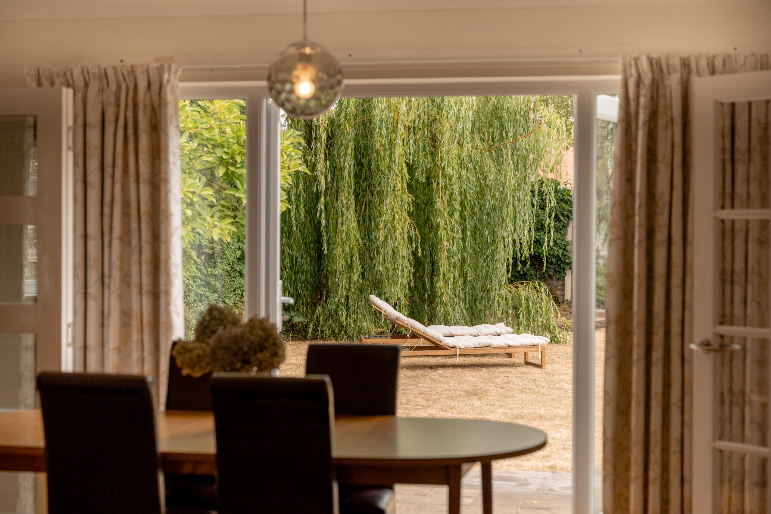 Dining room view through open doors to a yard with a lounge chair on dry grass, and a large willow tree in the background. A round light hangs above the dining table.
