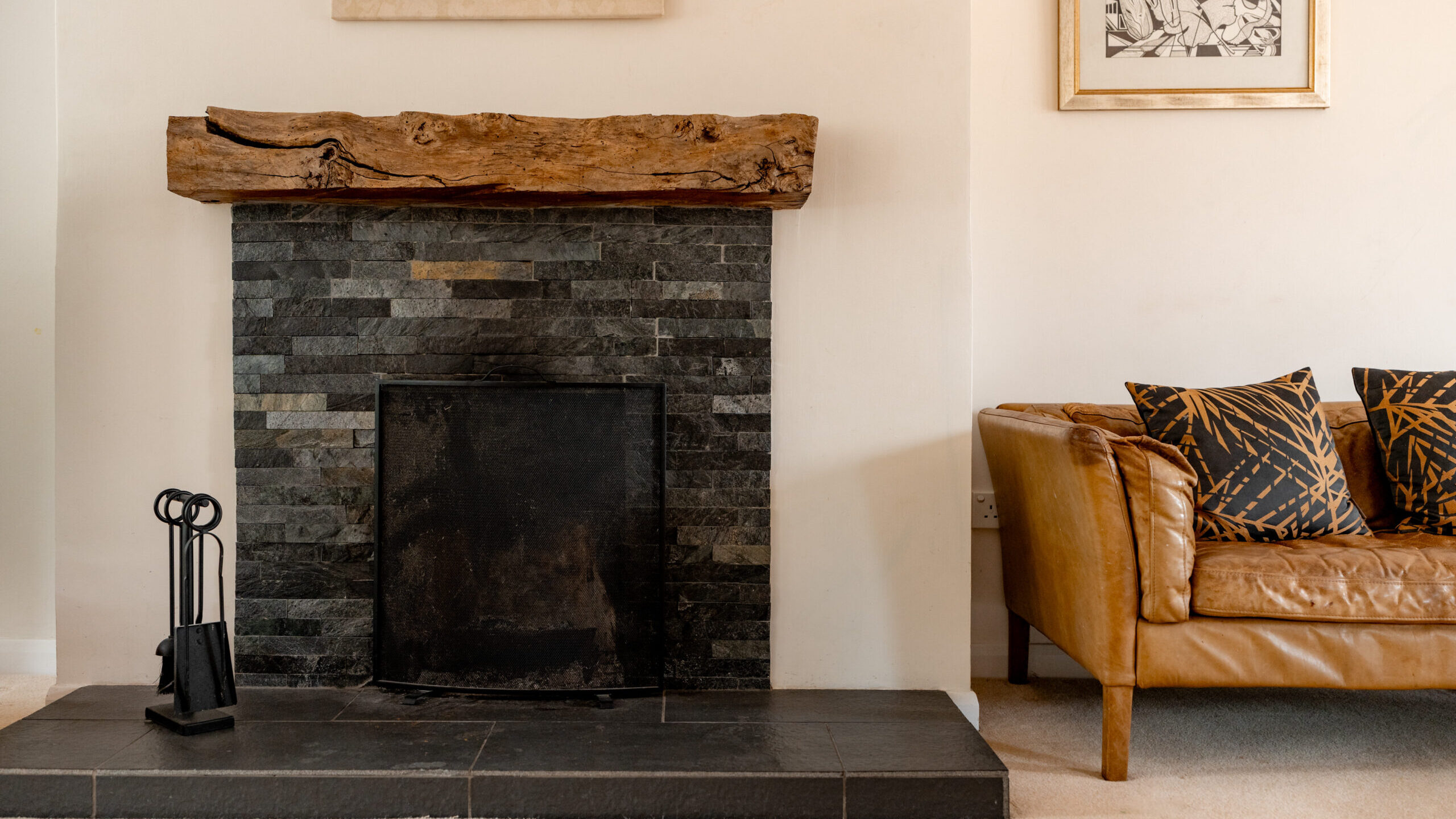 A modern living room with a black tiled fireplace, a rustic wooden mantel, fireplace tools, and a tan leather sofa with patterned cushions. Art hangs above the mantel and beside the sofa.