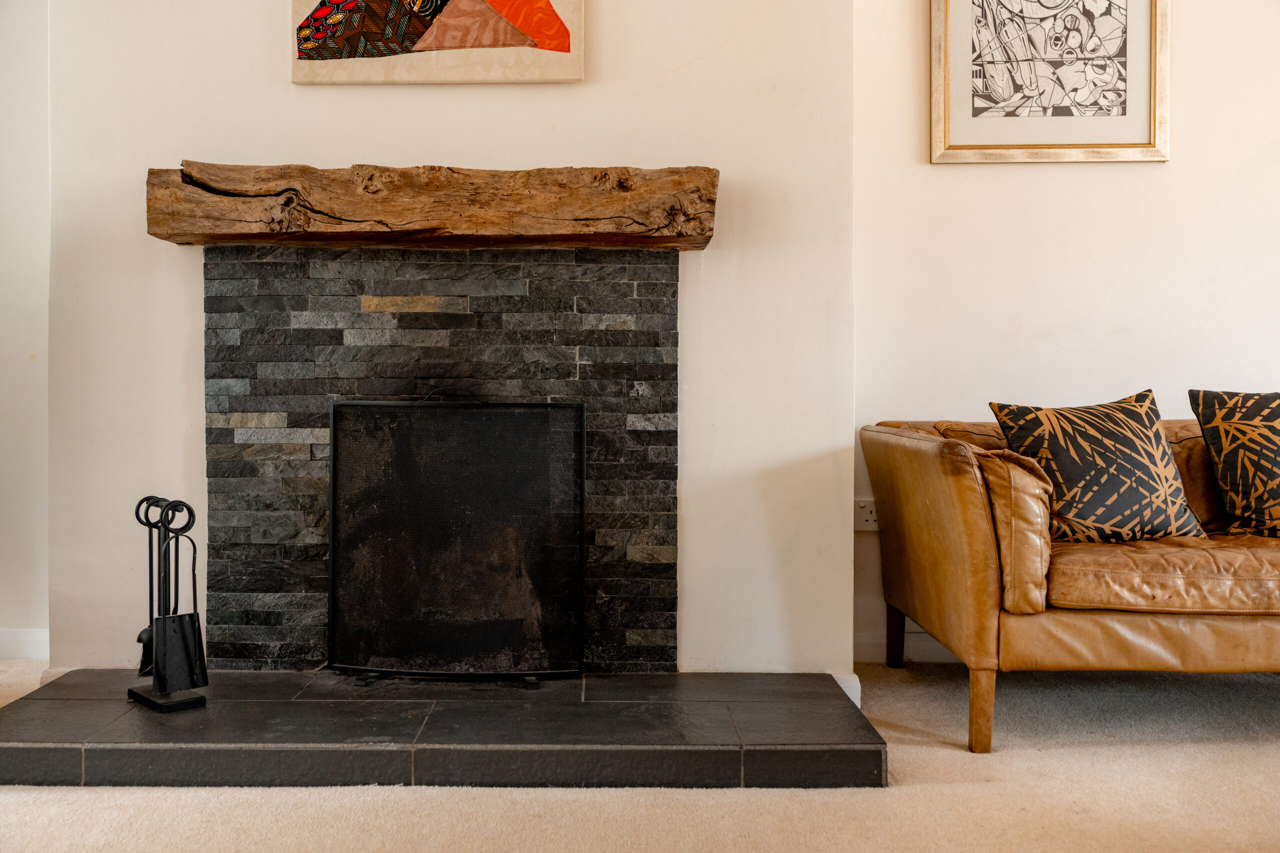 A modern living room with a black tiled fireplace, a rustic wooden mantel, fireplace tools, and a tan leather sofa with patterned cushions. Art hangs above the mantel and beside the sofa.