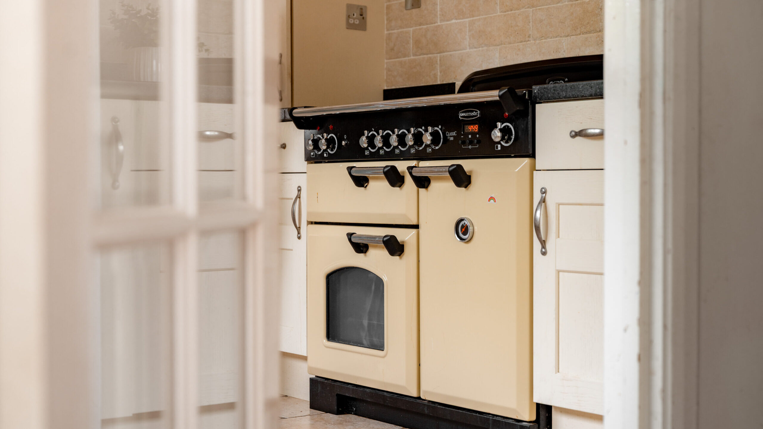 A cream-colored vintage oven with black knobs and handles is set against white cabinets and a beige tiled backsplash in a kitchen, viewed through a partially open door.