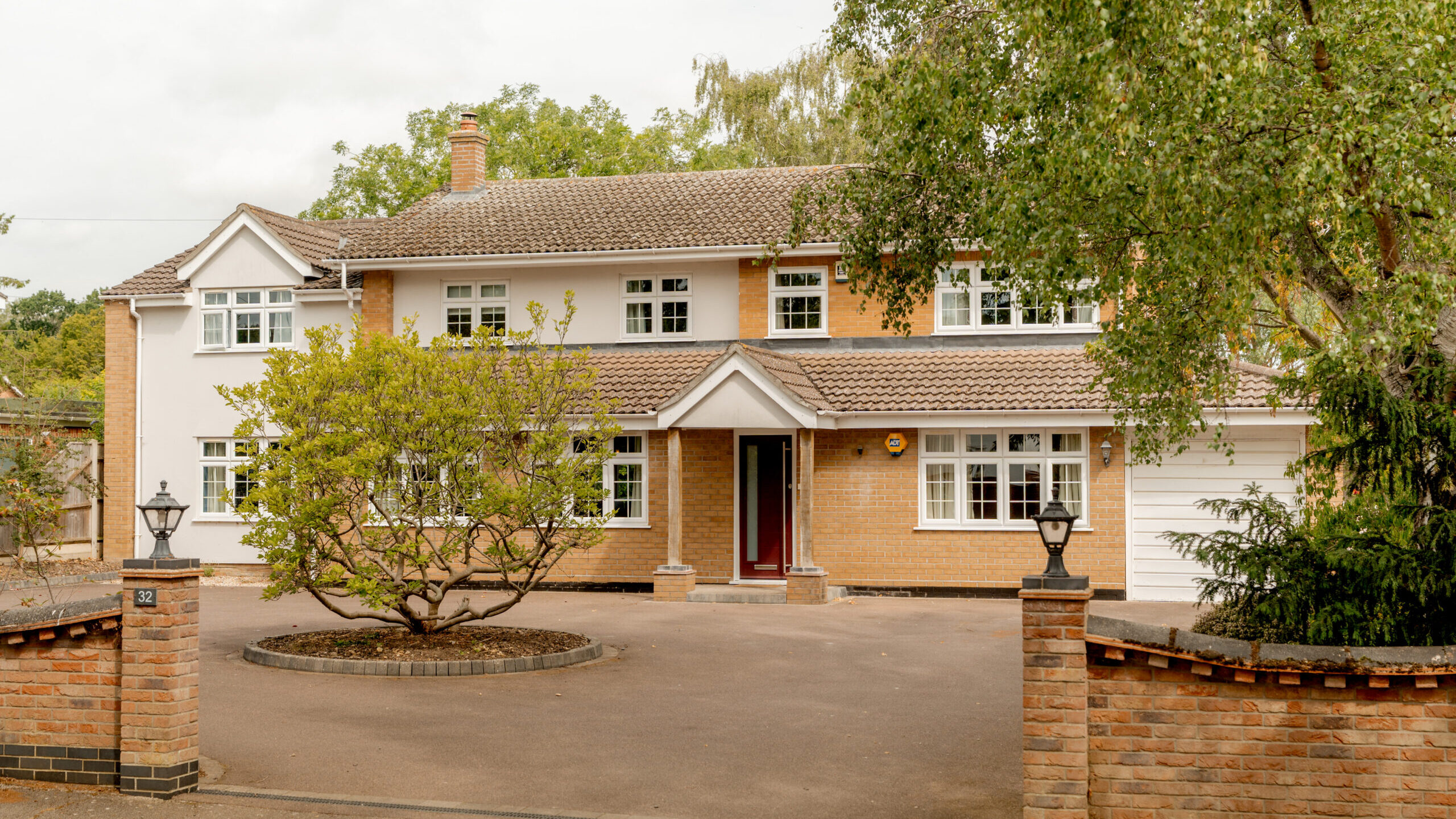 Two-story brick and beige house with a red front door, white trim, and large windows, surrounded by trees and a brick fence with lanterns. The driveway is spacious and clean.