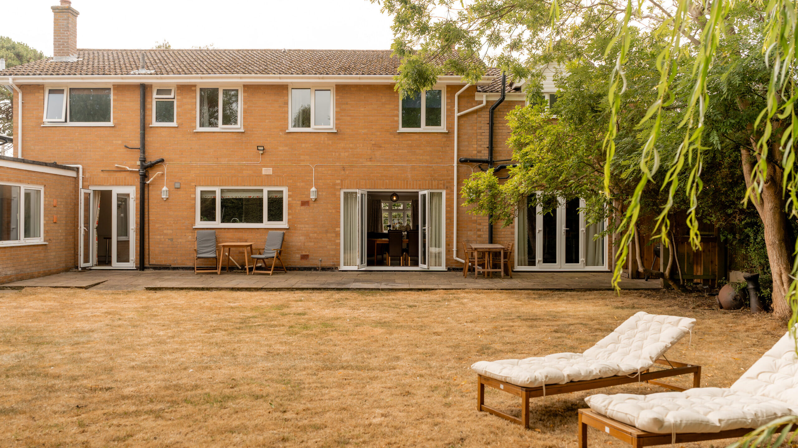 A two-story brick house with large windows and patio doors, a dry brown lawn, two white cushioned lounge chairs, and outdoor tables under leafy trees in the backyard.