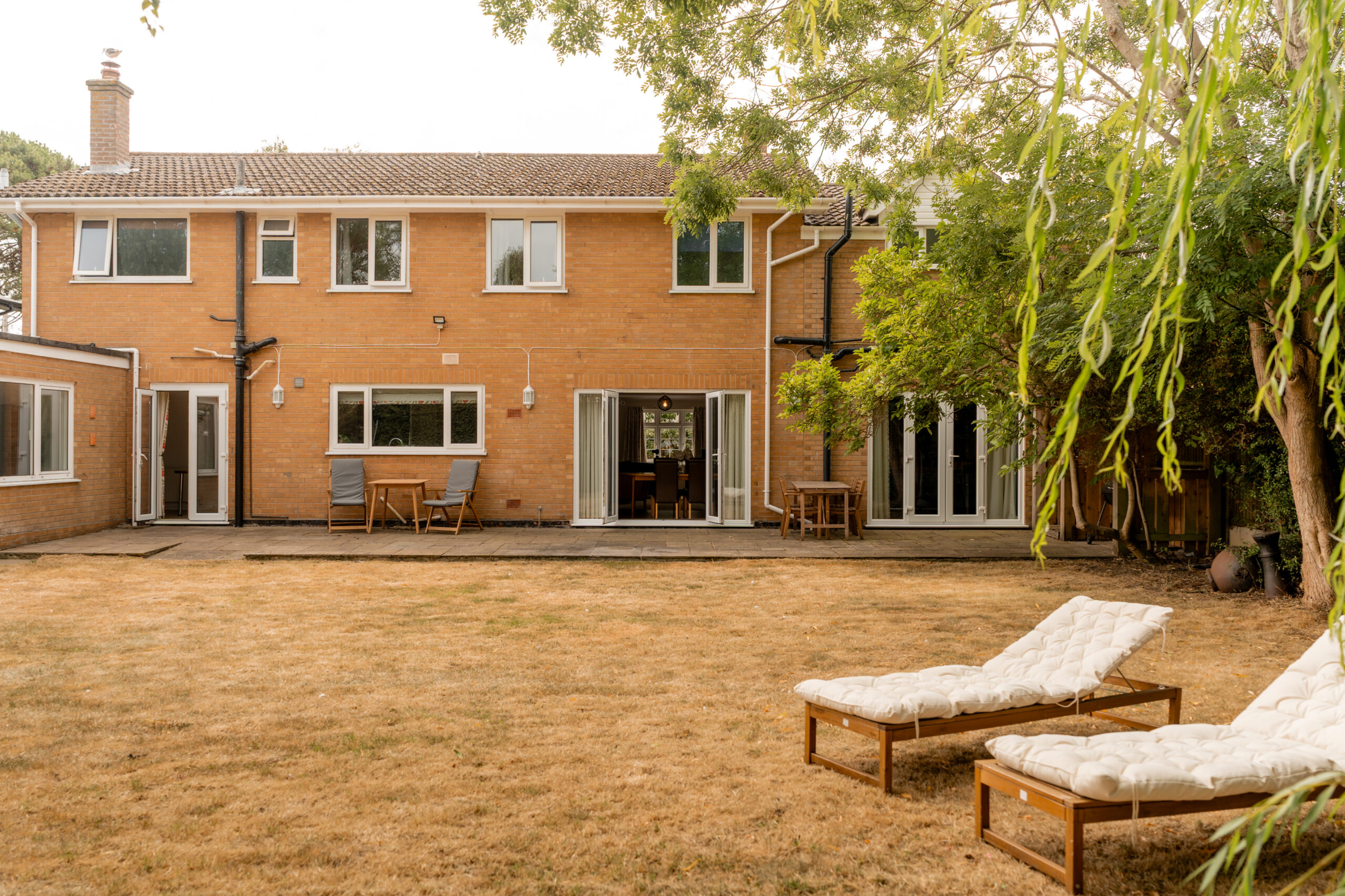 A two-story brick house with large windows and patio doors, a dry brown lawn, two white cushioned lounge chairs, and outdoor tables under leafy trees in the backyard.