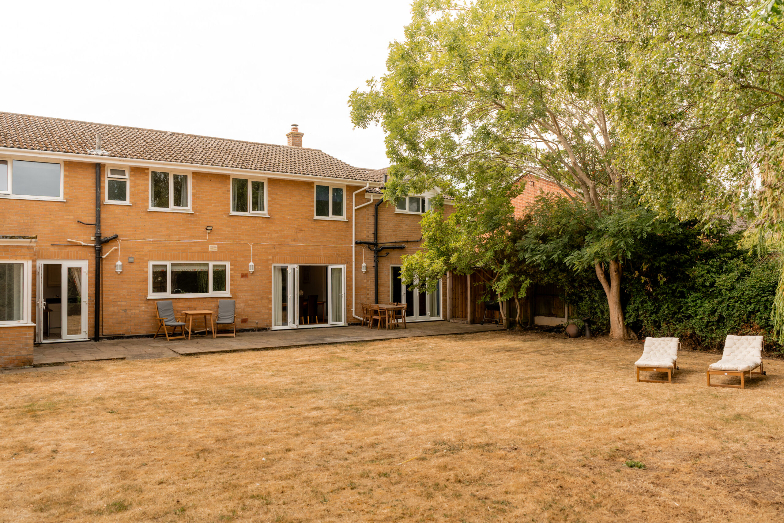 A two-story brick house with large windows and patio doors, a paved patio area with outdoor furniture, and a dry grassy backyard featuring two lounge chairs and several trees.
