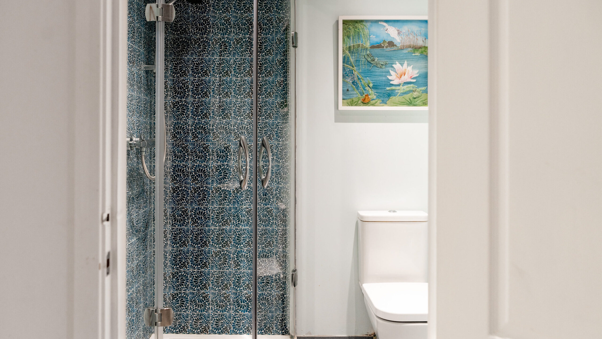 A bathroom with a glass shower featuring blue patterned tiles, a white toilet, and a framed painting of a water lily on the wall.