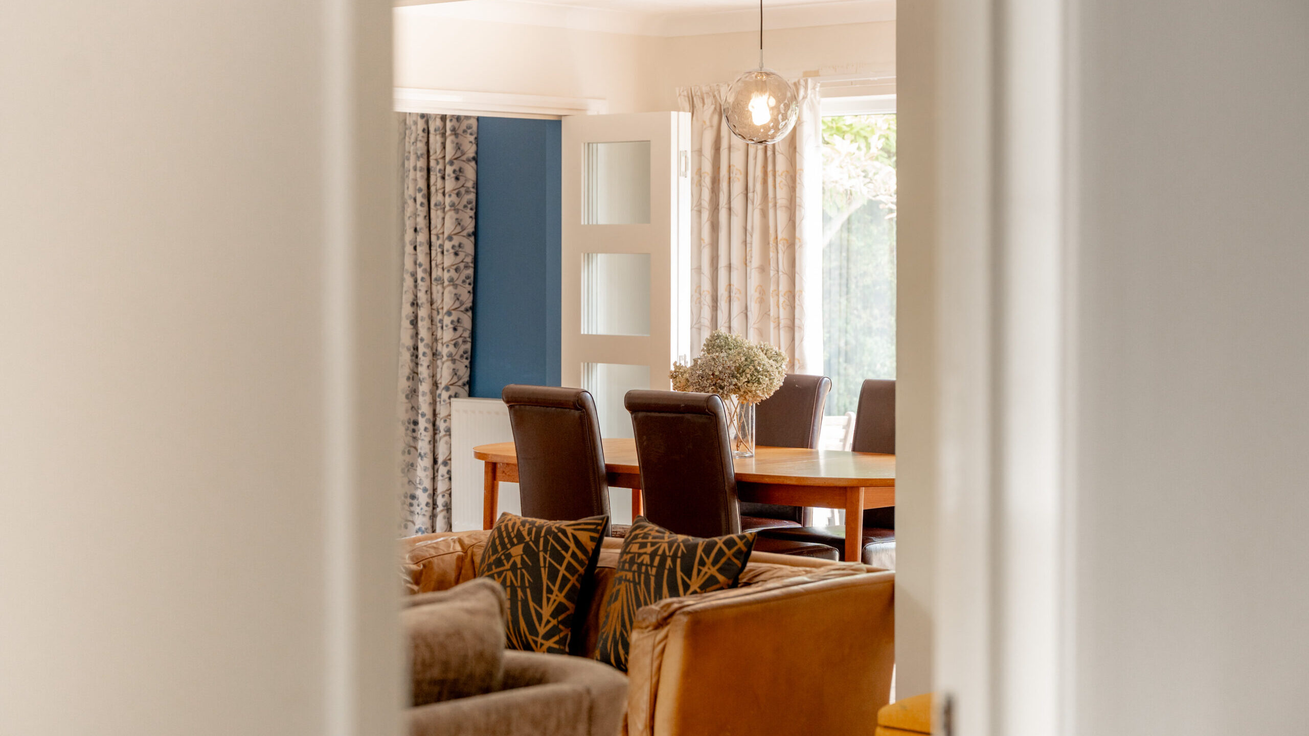 A cozy dining room with a wooden table, six brown chairs, a hanging light, and sunlight coming through large windows with patterned curtains, viewed through a doorway.