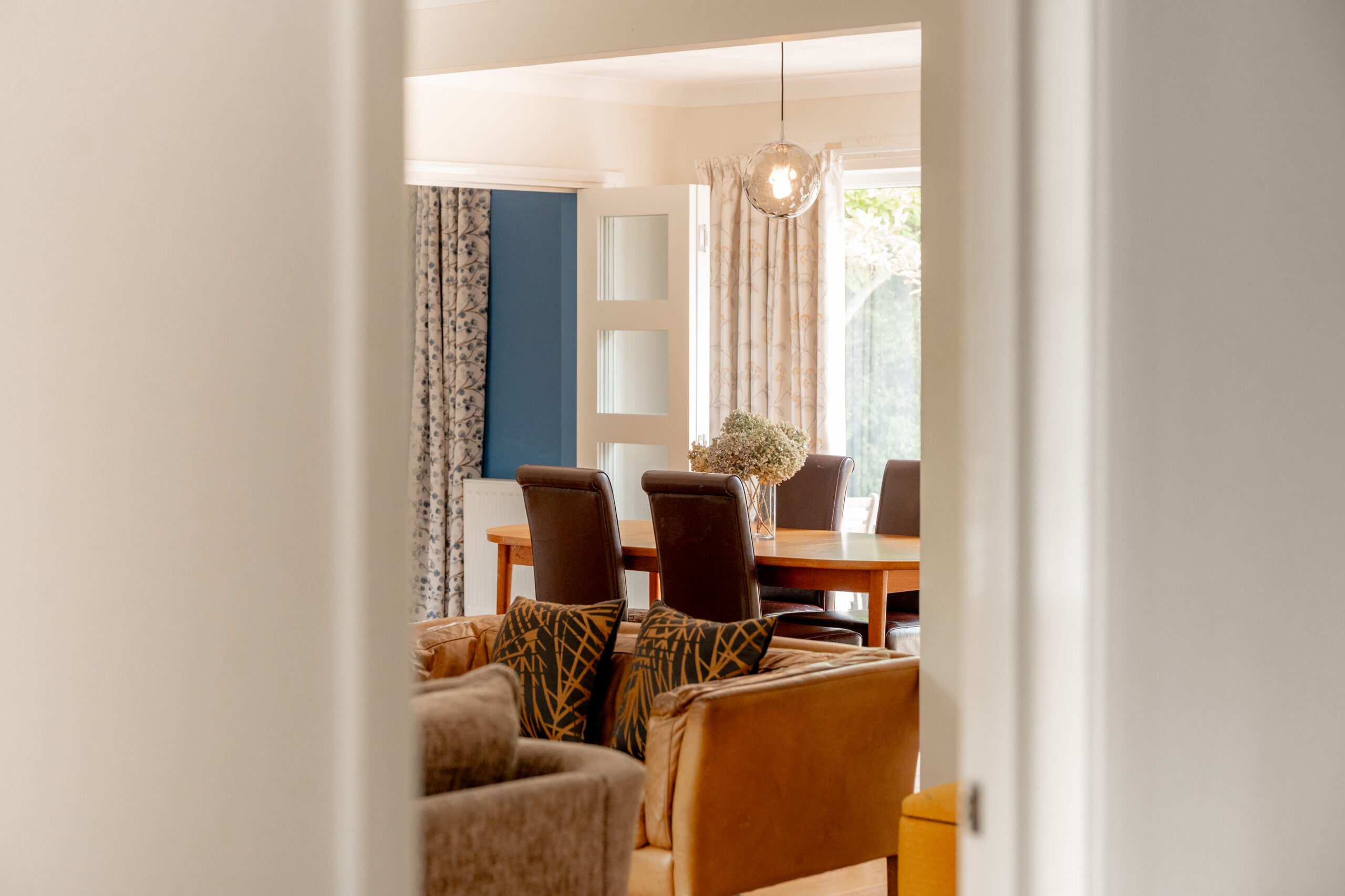 A cozy dining room with a wooden table, six brown chairs, a hanging light, and sunlight coming through large windows with patterned curtains, viewed through a doorway.