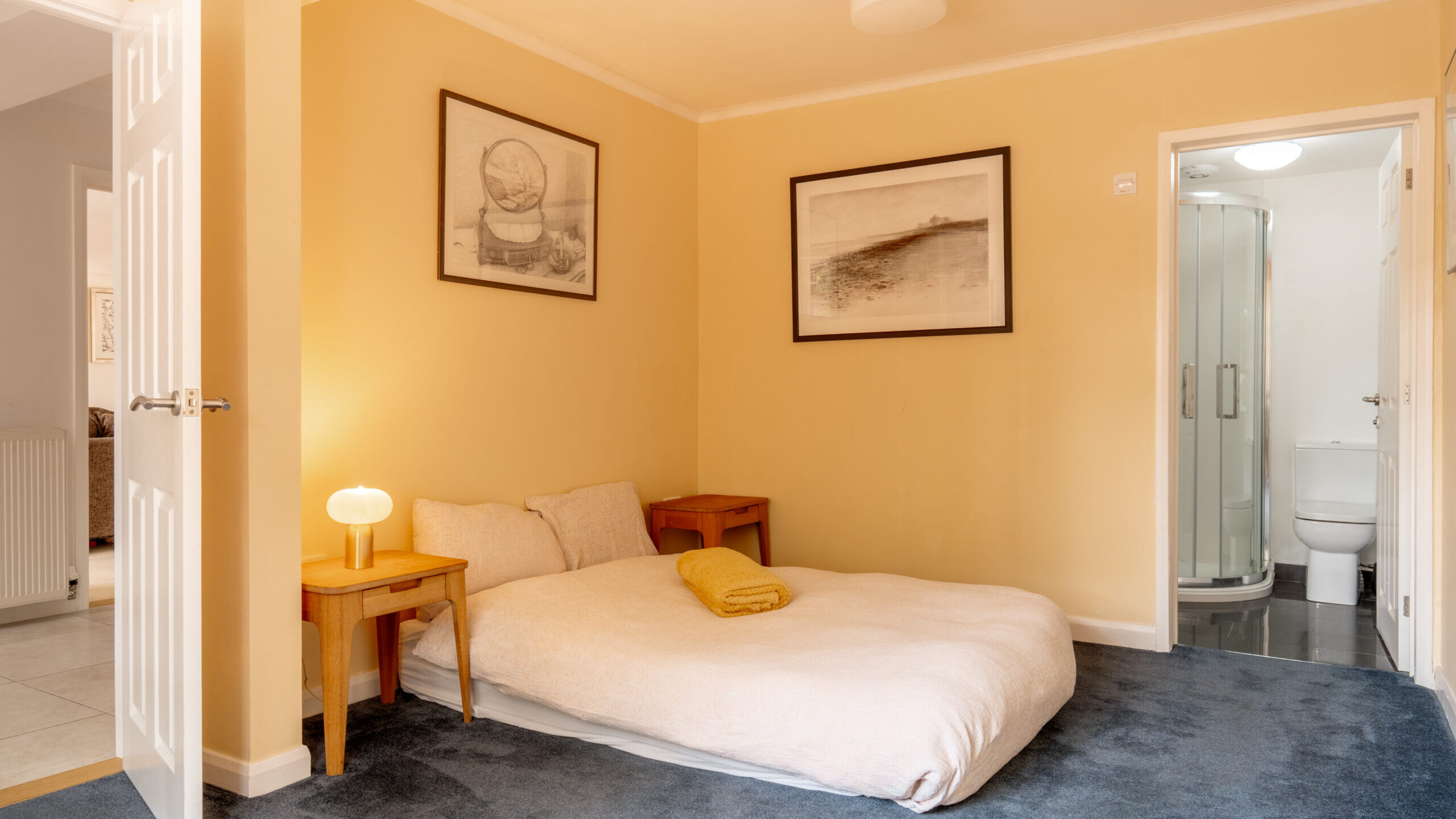 A cozy bedroom with a low double bed on blue carpet, two small wooden nightstands, framed art on yellow walls, a lamp, and an en-suite bathroom with a shower and toilet.