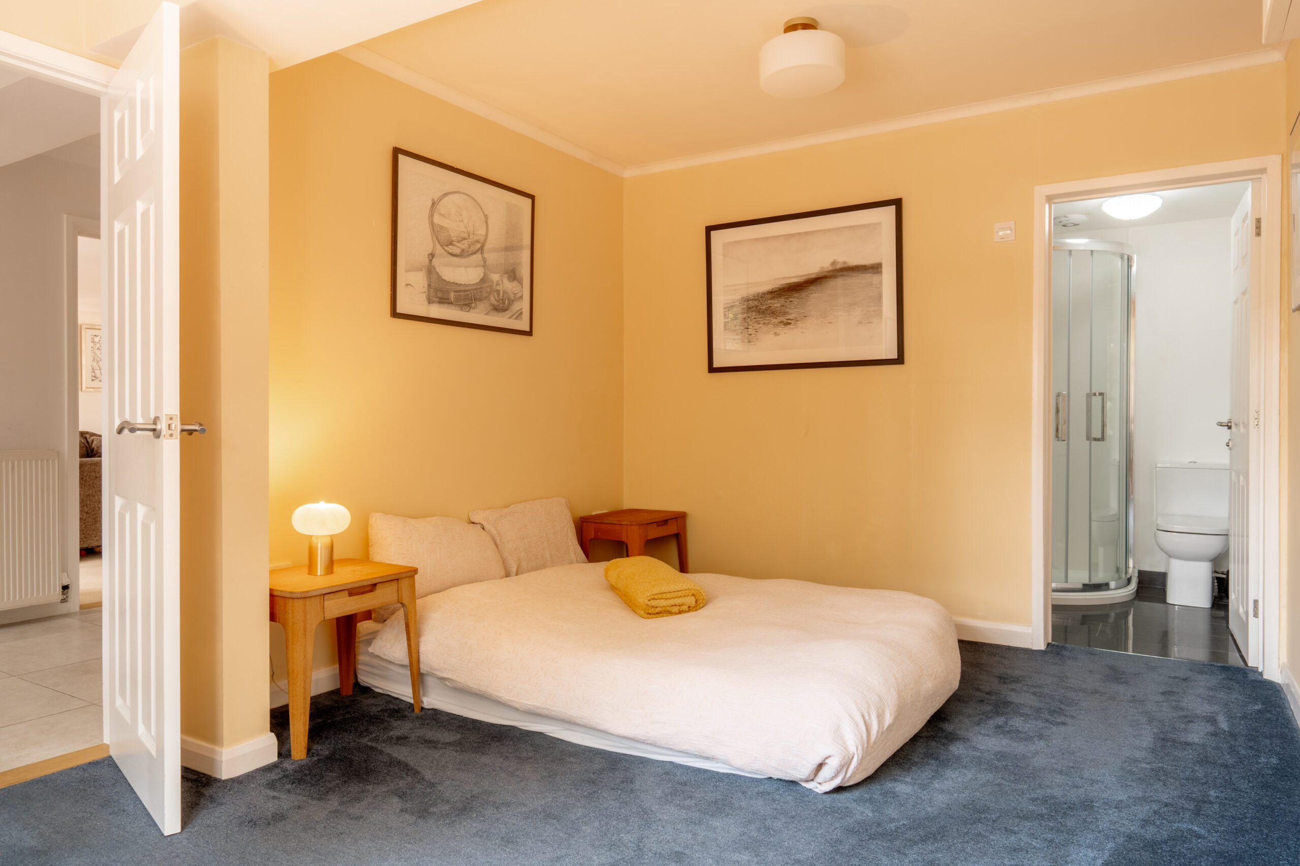 A cozy bedroom with a low double bed on blue carpet, two small wooden nightstands, framed art on yellow walls, a lamp, and an en-suite bathroom with a shower and toilet.