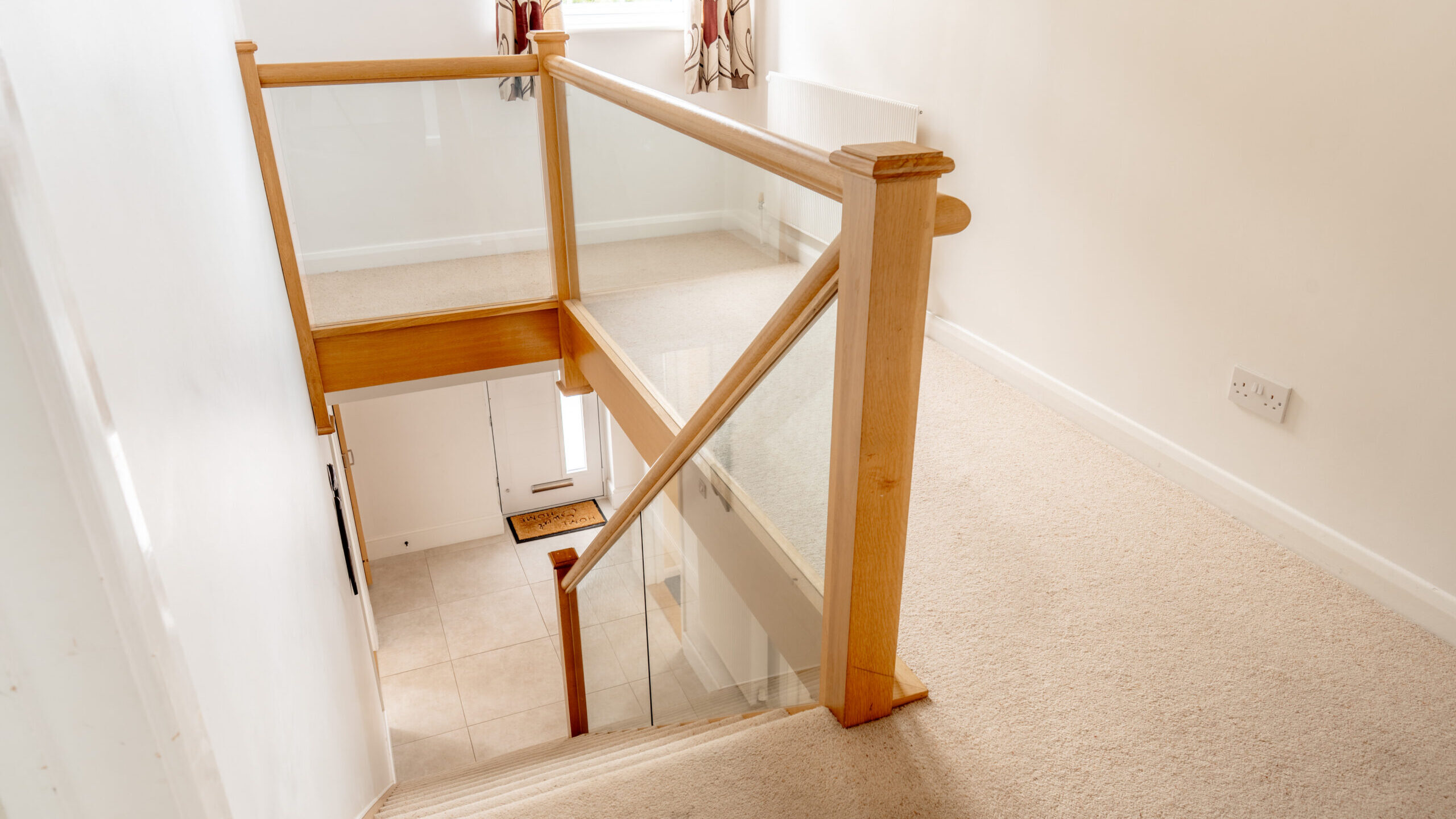 Bright hallway with beige carpet, wooden and glass railing, stairs leading down to a tiled entryway, white walls, radiator, two windows with patterned curtains, and a framed mirror on the wall.