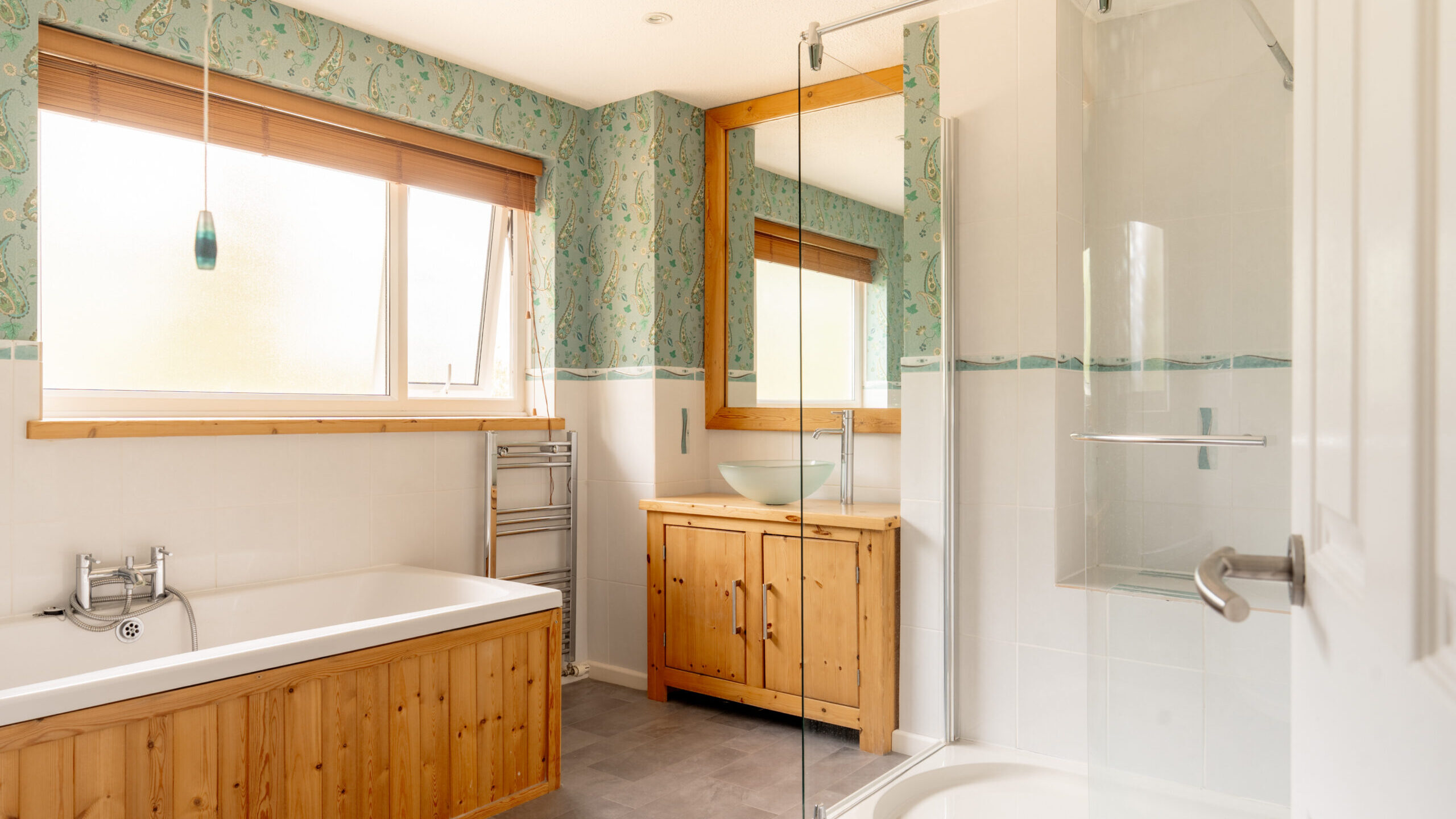 Bright bathroom with a wooden bathtub panel, glass shower, large window with wooden blinds, wooden vanity with a vessel sink, large mirror, and patterned wallpaper above white tiles.