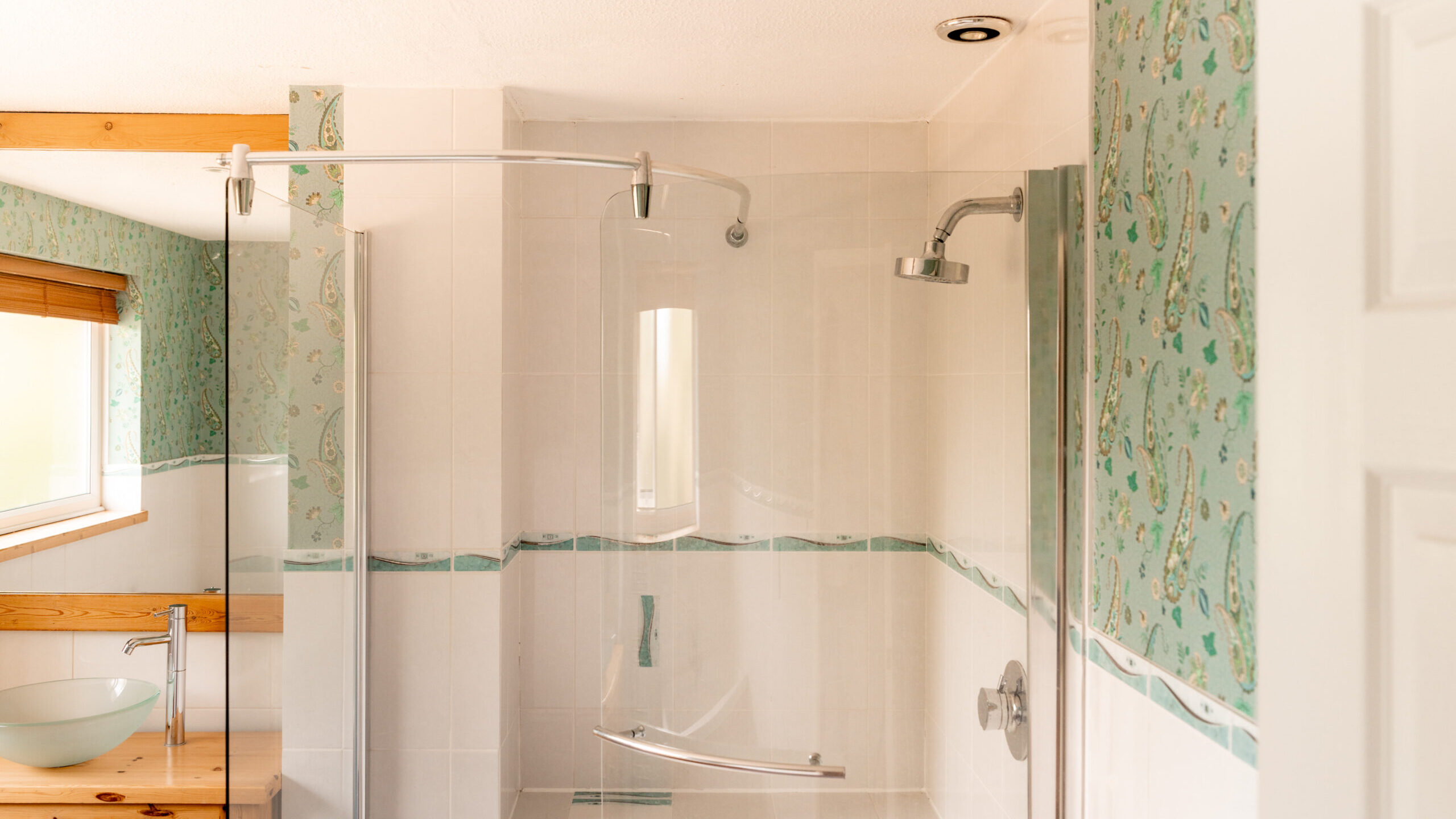 A bathroom with a walk-in shower, a glass door, and white tile. The walls have green floral wallpaper, and there is a wooden vanity with a glass vessel sink. A window with blinds is on the left.
