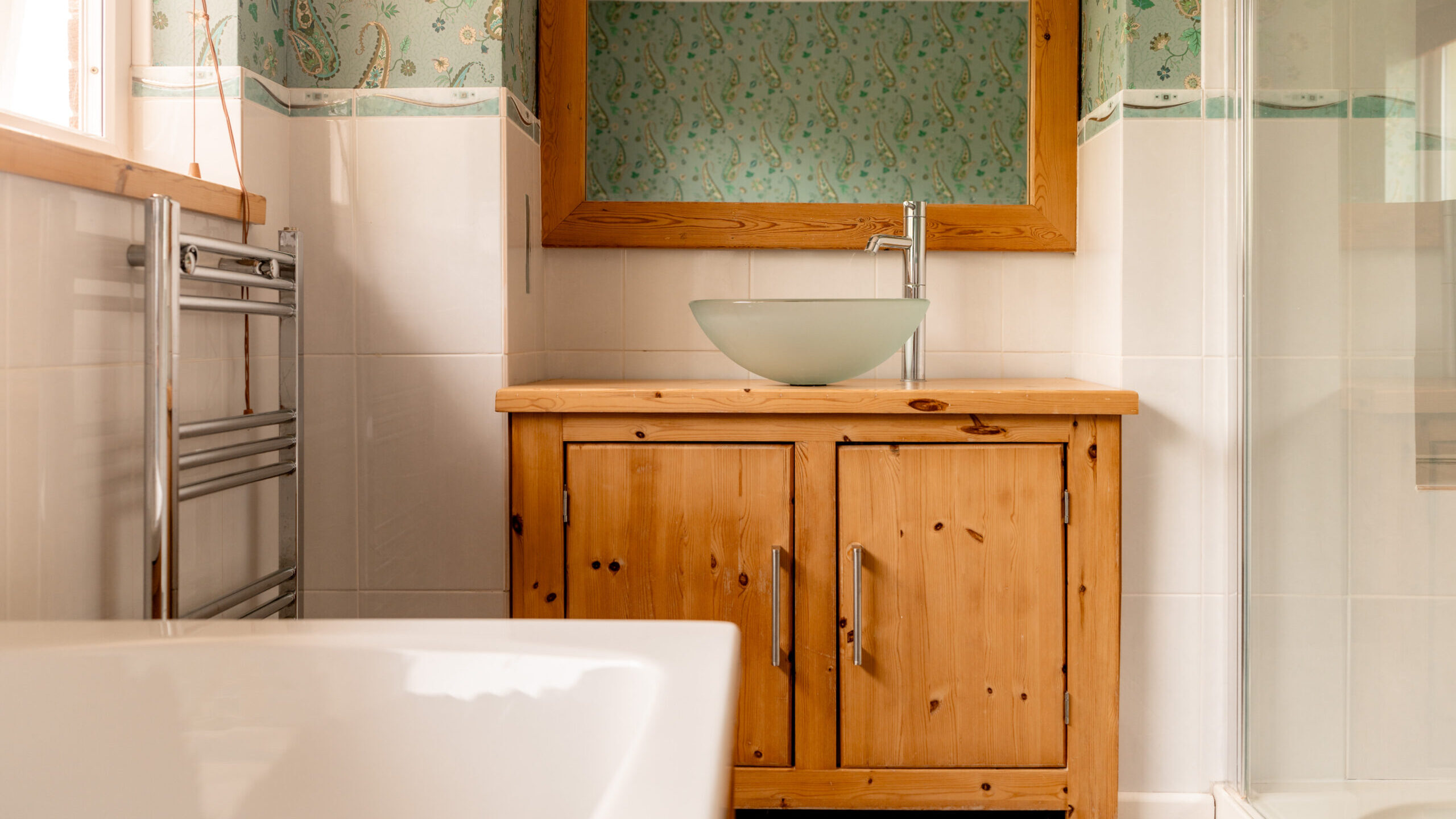 A bathroom with a wooden vanity, vessel sink, large mirror, white bathtub, glass shower, and patterned green wallpaper above white tiles.