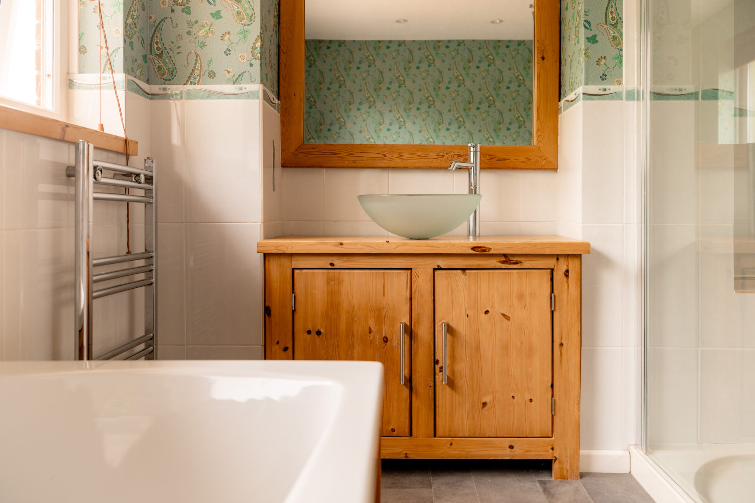 A bathroom with a wooden vanity, vessel sink, large mirror, white bathtub, glass shower, and patterned green wallpaper above white tiles.