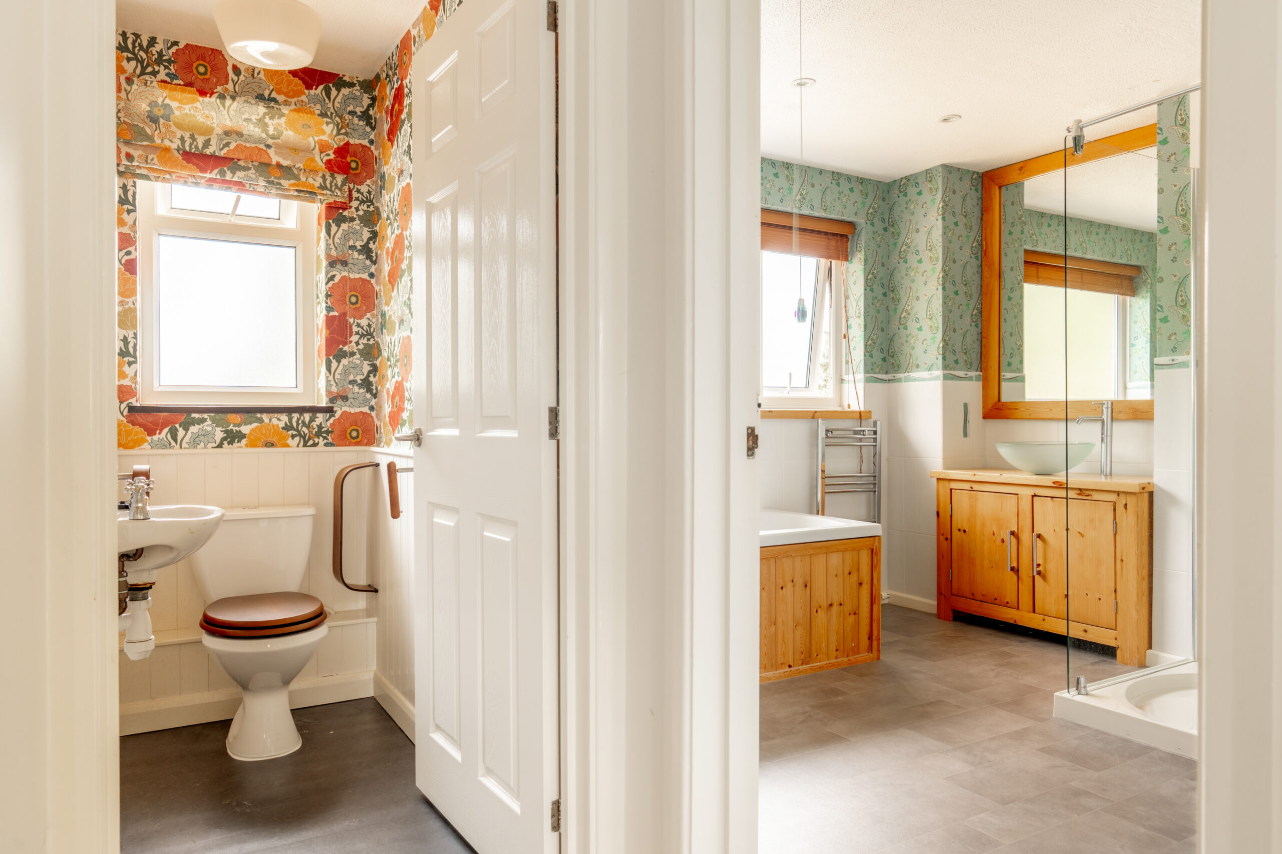 A view of two connected bathrooms, one with floral wallpaper, a toilet, and a grab bar, and the other with green wallpaper, a bathtub, shower, large mirror, and wooden vanity.