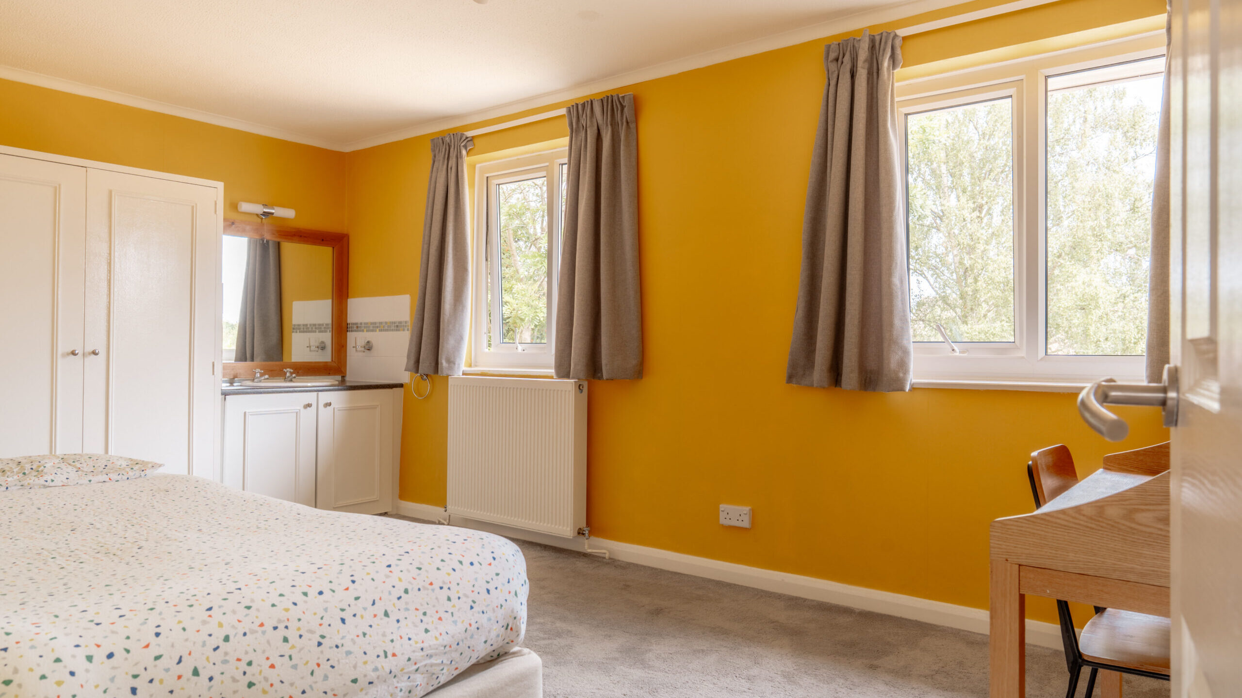 Bright bedroom with yellow walls, two windows with gray curtains, a bed with a dotted cover, a small vanity with a mirror, a closet, gray carpet, and a wooden desk with a chair.