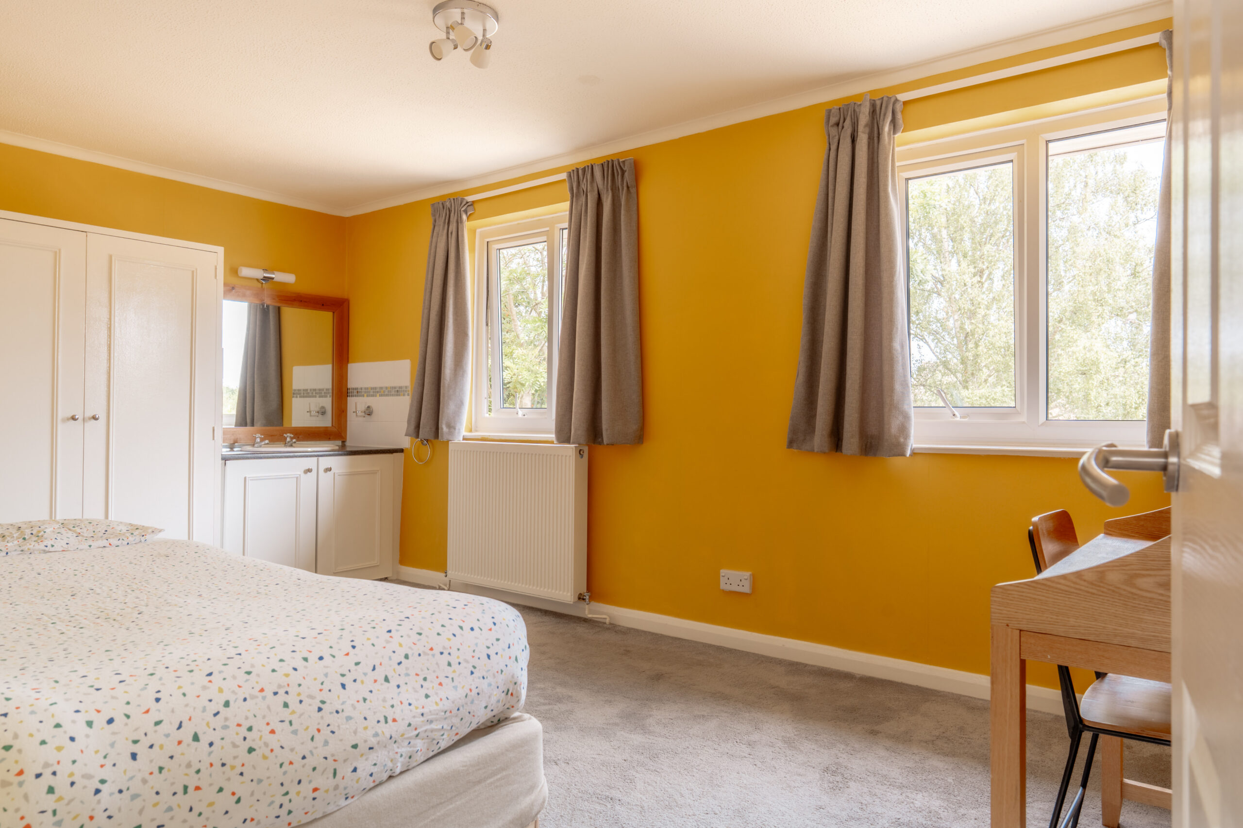 Bright bedroom with yellow walls, two windows with gray curtains, a bed with a dotted cover, a small vanity with a mirror, a closet, gray carpet, and a wooden desk with a chair.