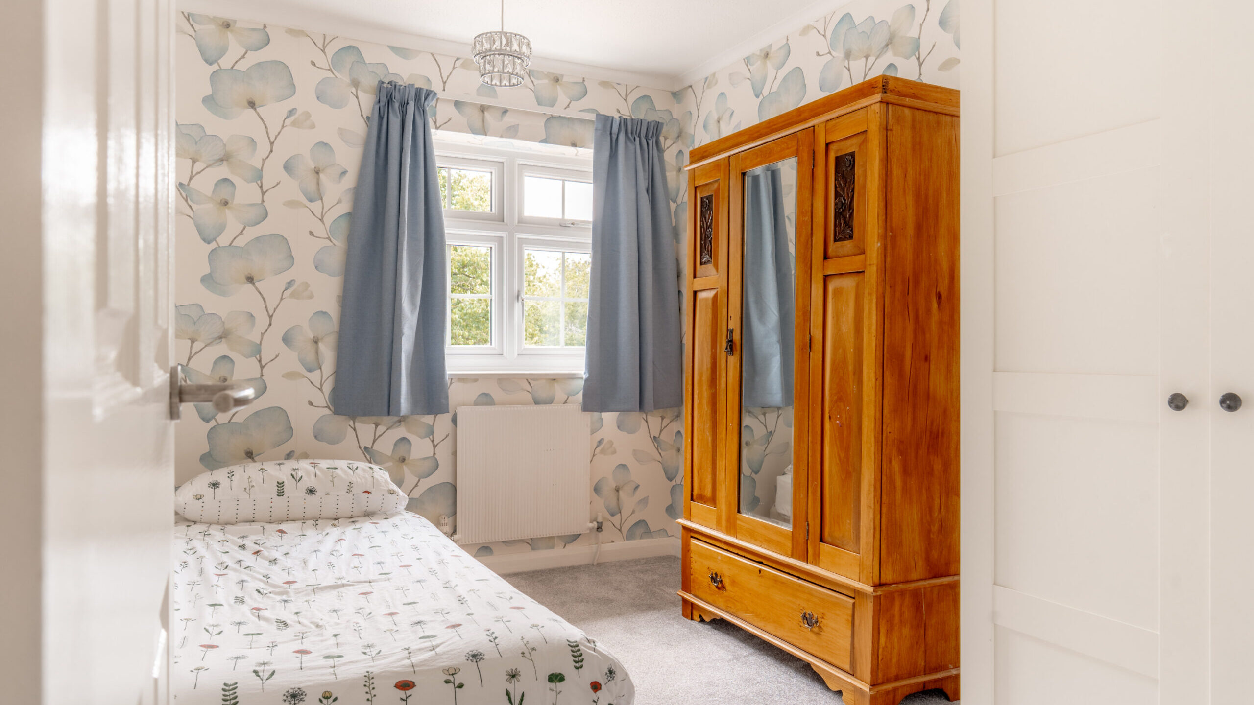 Small bedroom with a single bed, floral bedding, wooden wardrobe with mirror, blue curtains, and floral wallpaper. Light enters through a window above the bed, brightening the cozy room.