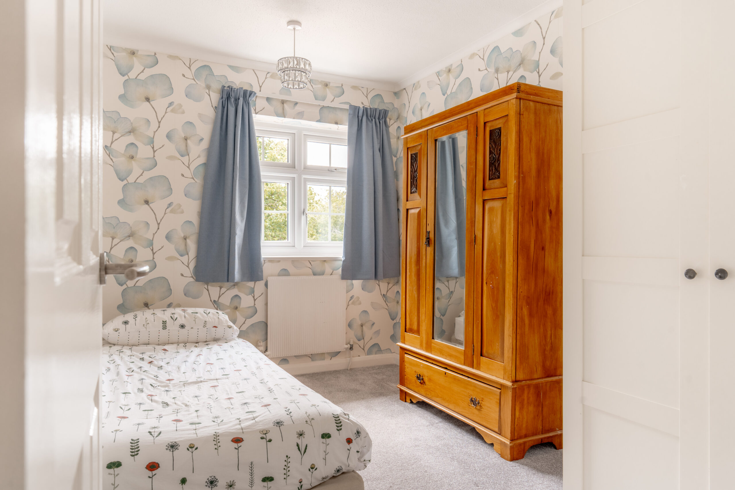 Small bedroom with a single bed, floral bedding, wooden wardrobe with mirror, blue curtains, and floral wallpaper. Light enters through a window above the bed, brightening the cozy room.
