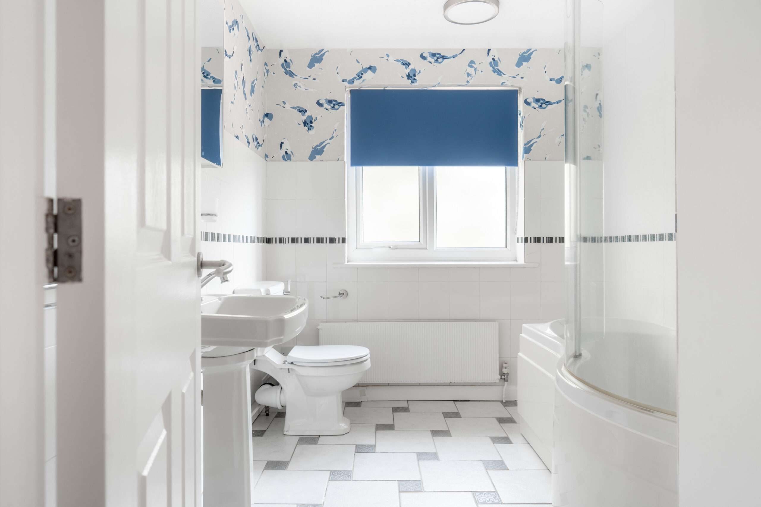 A bright bathroom with white fixtures, blue and white wallpaper, a bathtub with a curved glass screen, and a window with a blue blind. The floor is tiled in a gray and white pattern.