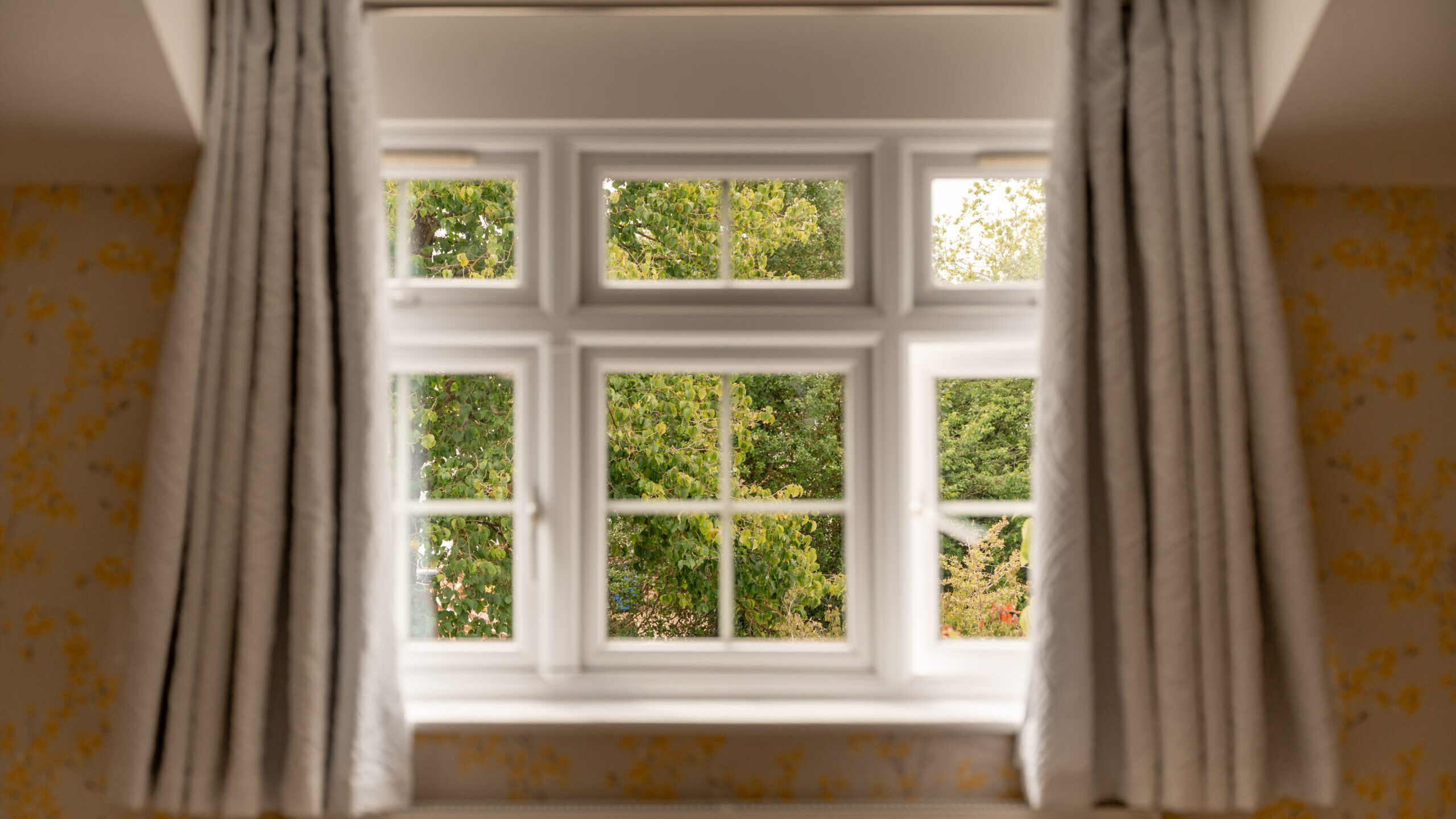 A white-paned window with beige curtains open on each side reveals a view of green trees outside. The window is letting in natural light, brightening the room.