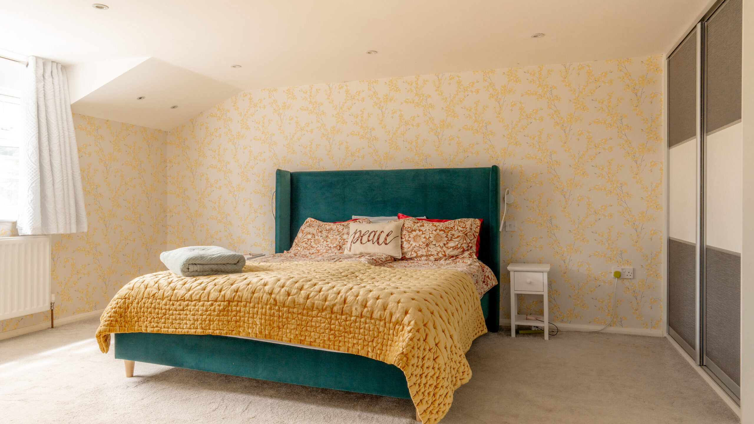 A bright bedroom with a green bed, mustard yellow blanket, patterned pillows, and a “peace” pillow. Yellow floral wallpaper, sliding wardrobe, white nightstand, and natural light from a window complete the cozy space.