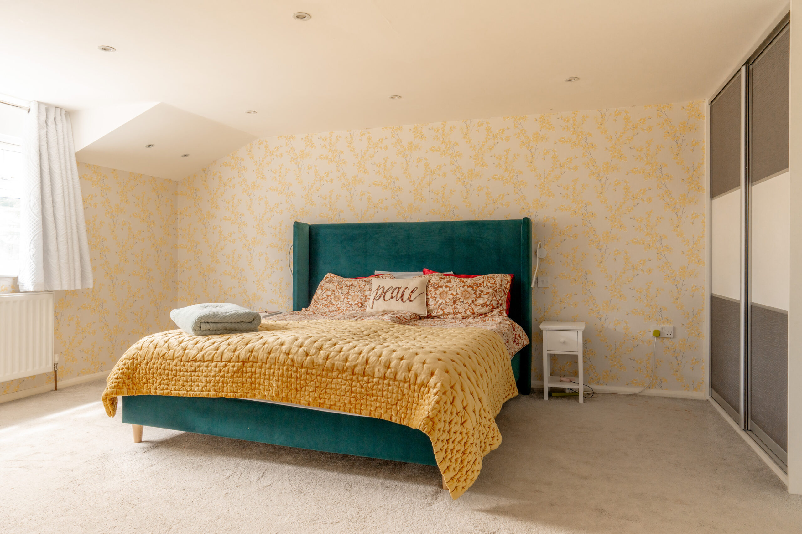 A bright bedroom with a green bed, mustard yellow blanket, patterned pillows, and a “peace” pillow. Yellow floral wallpaper, sliding wardrobe, white nightstand, and natural light from a window complete the cozy space.