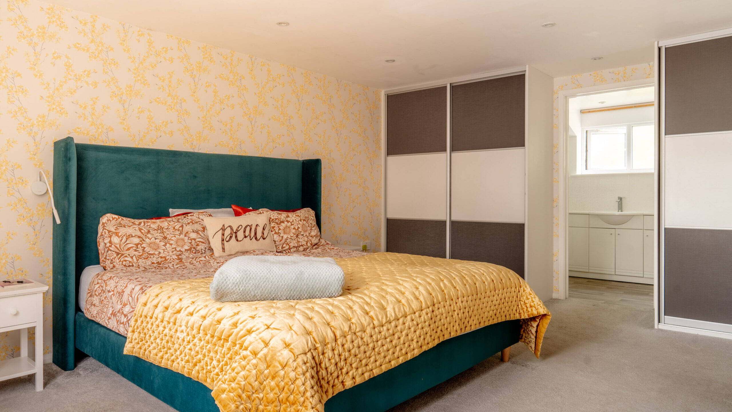 A bedroom with a teal bed frame, yellow patterned bedding, and a “peace” pillow. There are sliding closet doors, floral wallpaper, a nightstand, and an open door leading to a bathroom with a sink.