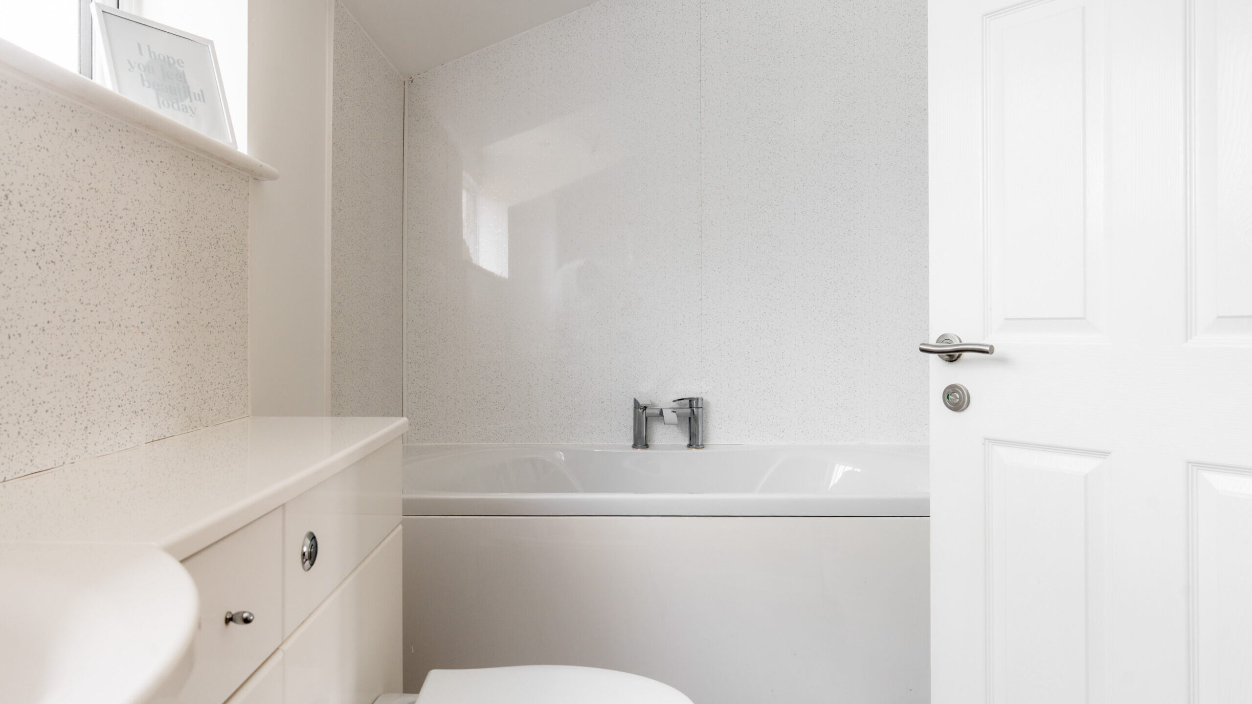 A modern white bathroom with a bathtub, toilet, and sink. The walls and cabinets are white, and there’s a faucet over the tub. Natural light enters through a window above the tub.