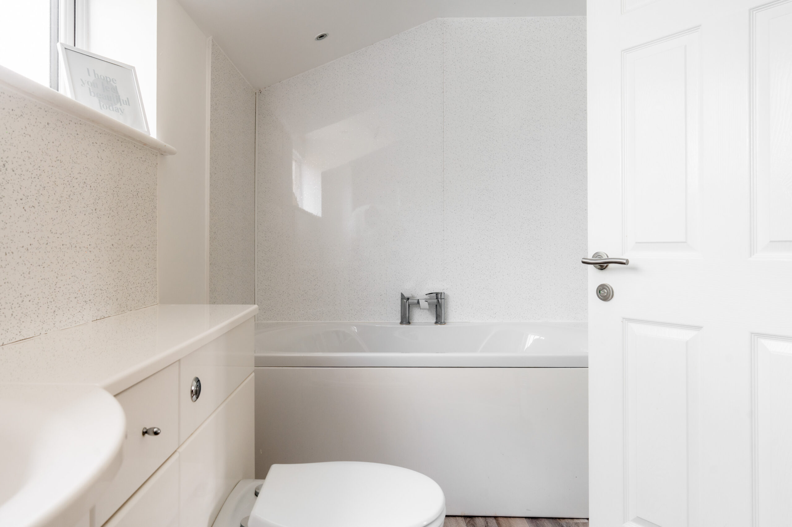 A modern white bathroom with a bathtub, toilet, and sink. The walls and cabinets are white, and there’s a faucet over the tub. Natural light enters through a window above the tub.