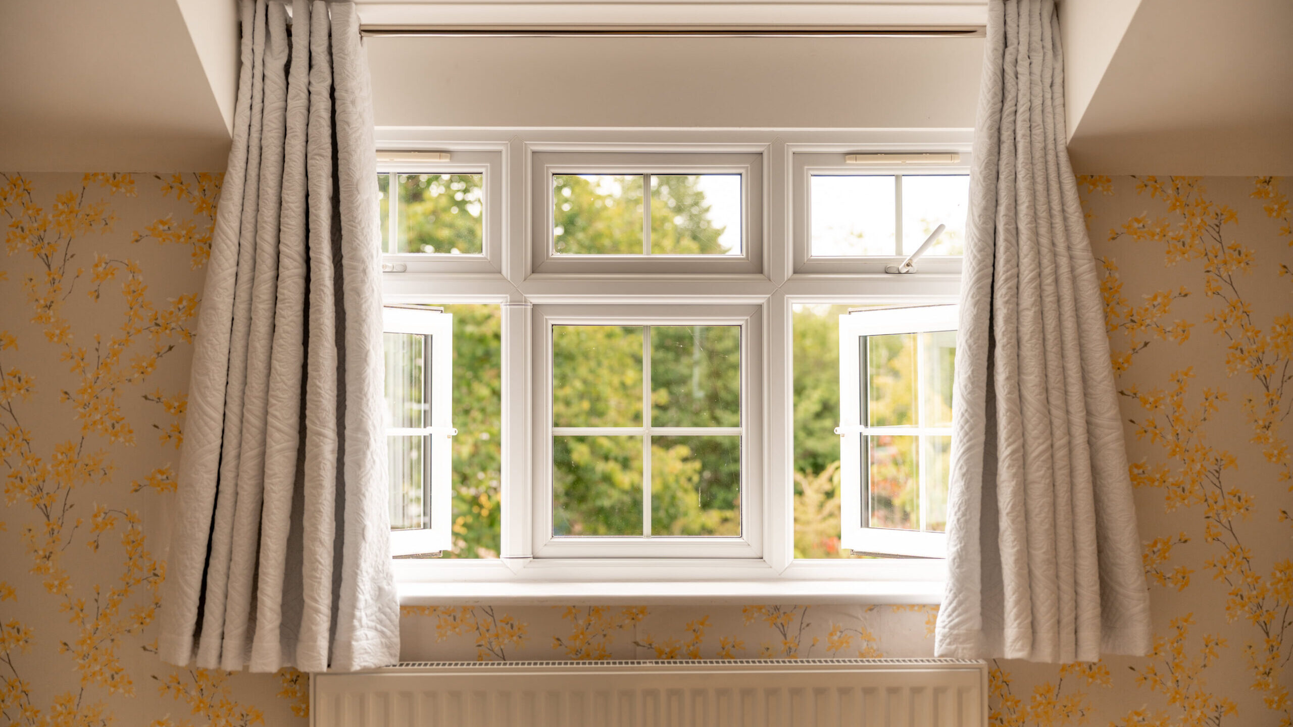 A white-framed window is open, with sheer white curtains on both sides. The window overlooks green trees and is set in a room with yellow floral wallpaper and a white radiator below.