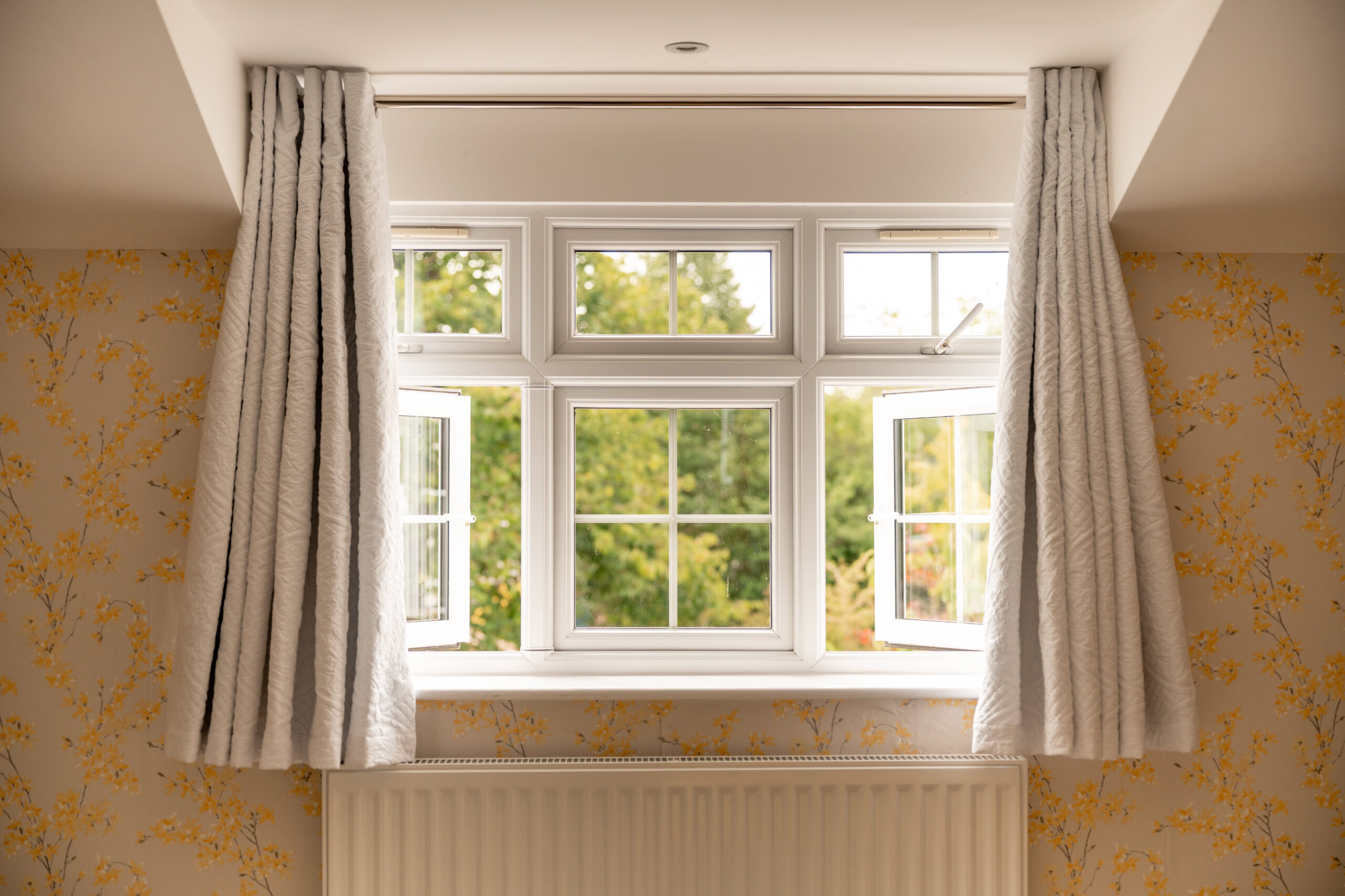 A white-framed window is open, with sheer white curtains on both sides. The window overlooks green trees and is set in a room with yellow floral wallpaper and a white radiator below.